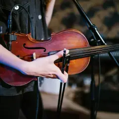 Dłoń muzyka trzyma smyczek nad skrzypcami. Ciepła barwa drewna instrumentu kontrastuje z ciemnym ubraniem.