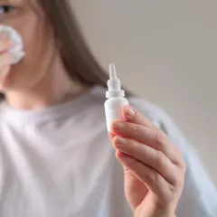 Kobieta z chusteczką przy nosie trzyma w dłoni małą buteleczkę z kroplomierzem.