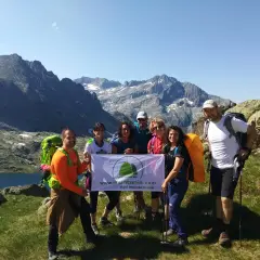Grupo de excursionistas sonríe en la montaña, listos para la ruta. Llevan mochilas y un cartel que dice "Aquí empieza tu viaje".