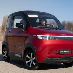 Czerwony, mały samochód elektryczny z kobietą za kierownicą, na tle placu i nieba.