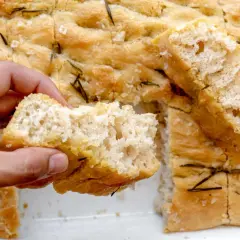 Mano rompe un trozo de focaccia original, dorada y con romero.