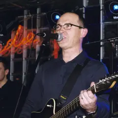 Wokalista z gitarą śpiewa na scenie, a w tle neonowy napis. Czas na koncert, a kiedy przyjdzie na ciebie czas tekst.