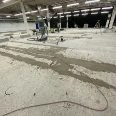 Prace przy naprawie posadzki betonowej. Widać sprzęt budowlany, pracowników i przygotowane do wylania fugi.