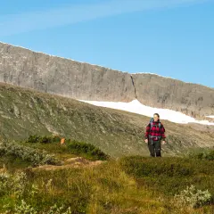 En kvinna på helags topptur njuter av utsikten över fjällmassivet. Solen lyser på den gröna sluttningen.