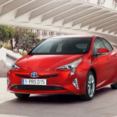 Czerwony Prius 4 z nowoczesnym designem, zaparkowany pod betonową konstrukcją z palmami w tle.
