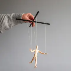 Dłoń w swetrze steruje drewnianym ludzikiem na sznurkach, symbolizując psychologię manipulacji i kontroli.