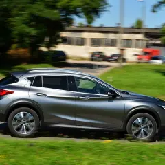 Srebrny Infiniti QX30 pędzi po drodze, mijając zielone pobocze i budynki.