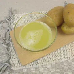 Szklanka z pianistym sokiem z ziemniaka stoi obok kilku surowych ziemniaków na drewnianej desce.