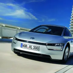 Srebrny Volkswagen XL1 sunie po drodze obok futurystycznego budynku.