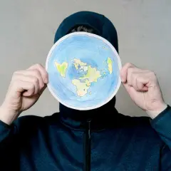 Osoba w kapturze trzyma okrągły rysunek płaskiej Ziemi. To nie jest globe po polsku, ale ciekawa wizja świata.