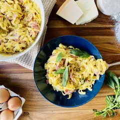 Un plat de pâtes à la carbonara crémeuses, garni de basilic frais. Des ingrédients comme des œufs, du fromage et de la farine sont visibles à proximité.