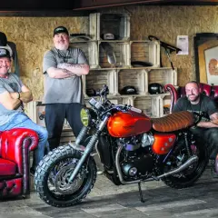 Three men pose with a custom orange Triumph motorcycle, showcasing the work of best custom bike builders.