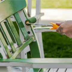 En hand med plasthandske målar en trädgårdsstol grön med en liten roller. Det är dags att måla snickerier utomhus.