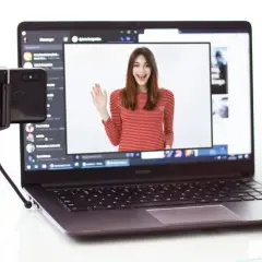 Kamera z telefonu na PC: młoda kobieta w bluzce w paski wita się na ekranie laptopa, obok stoi telefon na statywie.