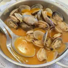 Almejas en una cazuela metálica, bañadas en una sabrosa salsa marinera. Una cuchara plateada reposa sobre el plato, lista para disfrutar.