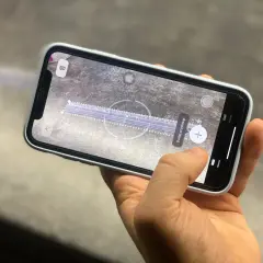 Dłoń trzyma smartfon, na ekranie którego działa aplikacja miarka. Wirtualna linijka mierzy odległość na powierzchni.