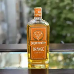 Butelka Jägermeister Orange, z pomarańczowym akcentem, stoi na szklanym blacie.