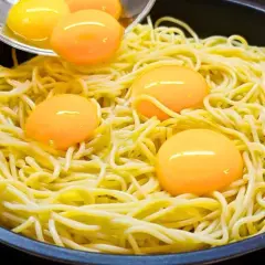 Makaron z jajkiem w patelni, żółtka tworzą złote kropki na żółtym tle.