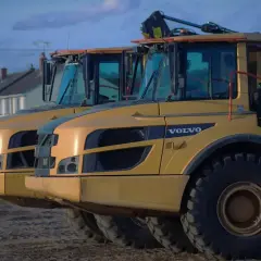 Dwa żółte wywrotki Volvo A30D stoją obok siebie. W tle domy i niebo.