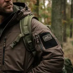 Mężczyzna w brązowej kurtce z naszywką Helikon-Tex, symbol polskie marki sportowe, w lesie.