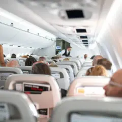 Kobieta w białej sukience stoi w przejściu samolotu, podczas gdy pasażerowie siedzą w rzędach. Widać lekkie turbulencje w samolocie.