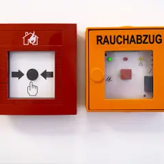 RWA Anlagen Wartung: Rote Brandmeldeanlage und orange Rauchabzugseinheit an weißer Wand.