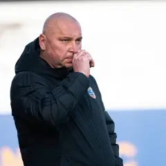 Mariusz Lewandowski, trener w czarnej kurtce, zamyślony, z ręką przy nosie.