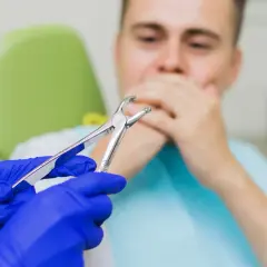 Dentysta w niebieskich rękawiczkach trzyma kleszcze. Pacjent zakrywa usta. Czy po wyrwaniu zęba można wychodzić na dwór?