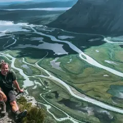 Man vilar på en klippa med utsikt över Sarek nationalparks meandrande vattendrag och fjällandskap.