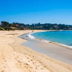 La **playa pintens** se extiende bajo un cielo azul intenso, con aguas turquesas acariciando la arena dorada. Casas y vegetación salpican la costa.