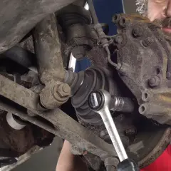 Mechanik w czerwonej koszulce z logo "Auto" dokonuje czyszczenia zacisków hamulcowych, używając klucza nasadowego.