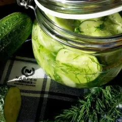 Burk med inlagd gurka och färsk dill. Bredvid står hela gurkor och en skiva.