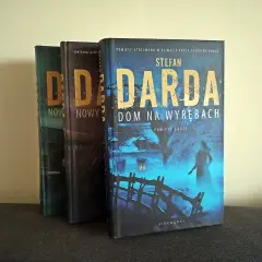 Trzy książki Stefana Dardy, w tym "Dom na wyrębach", stoją obok siebie. Okładka "Domu na wyrębach" przedstawia mroczny, niebieski krajobraz z domem i postacią.