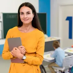 Nauczycielka w pomarańczowej sukience trzyma tablet w klasie, gdzie dzieci siedzą przy ławkach.