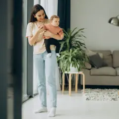 Uśmiechnięta kobieta z dzieckiem na rękach stoi przy oknie w jasnym salonie.