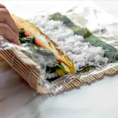 Ręce zwijają domowe sushi na macie bambusowej. Ryż, nori, omlet i warzywa tworzą pyszne wnętrze rolki.