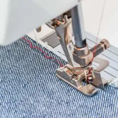 Sewing machine foot guiding denim fabric, demonstrating how to blind hem pants with a red thread stitch.