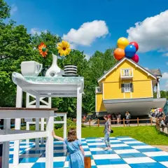 Dzieci bawią się w surrealistycznym parku, gdzie dom unosi się na balonach, a stół jest gigantyczny. Idealne miejsce na rodzinne przygody w parkach dla dzieci.
