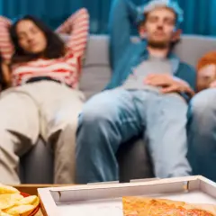 Grupa przyjaciół odpoczywa na kanapie po obfitym posiłku, czując senność. Na stole pizza, chipsy i piwo.