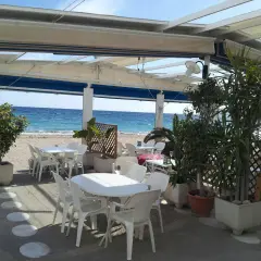 Terraza de restaurante en la playa. Mesas y sillas blancas bajo un toldo, con el mar azul de fondo. Un lugar perfecto para comer con agua amarga.