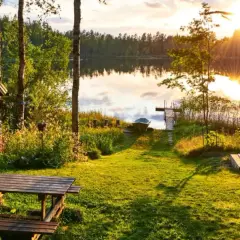 Letnia chata nad jeziorem w Finlandii. Słońce zachodzi, rzucając złote światło na trawę i wodę. Ceny w Finlandii mogą być wysokie, ale widoki są bezcenne.