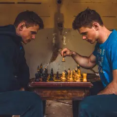 Dwóch młodych mężczyzn gra w szachy w opuszczonym miejscu. Jeden z nich, w niebieskiej koszulce, właśnie wykonuje ruch pionkiem, pokazując, jak bije pionek w szachach.