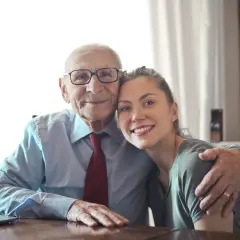 Uśmiechnięty starszy pan w okularach i młoda kobieta przytulają się. Widać ich twarze i ramiona. To piękny obraz łączący pokolenia.