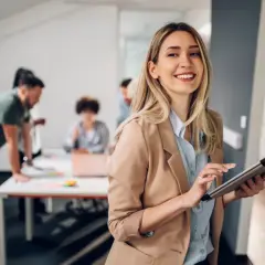A smiling woman with a tablet, likely pursuing her masters in leadership and management, stands in an office as colleagues collaborate in the background.