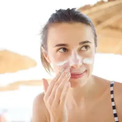 Frau trägt Sonnencreme auf, um Pickel durch Sonne zu vermeiden. Sie ist am Strand unter Sonnenschirmen.
