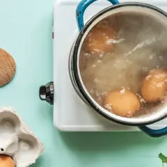 Des œufs sont en train de cuire des œufs durs dans une casserole d'eau bouillante. Une cuillère en bois et une boîte à œufs vide sont à proximité.