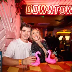 Coppia sorridente in un locale di Londra, con cocktail rosa e un'insegna al neon "DOWNTOWN".