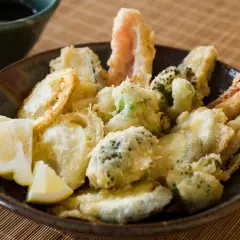 A bowl of crispy vegetable tempura, featuring broccoli, zucchini, and shrimp, served with lemon wedges and a side of dipping sauce.