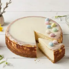 Pyszny sernik na Wielkanoc, udekorowany pastelowymi jajeczkami i lukrem. Obok słoiczek z konfiturą malinową i wiosenne kwiaty.