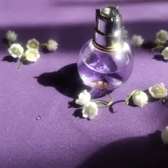 Szklana buteleczka perfum z fioletowym płynem i konwalią na fioletowym tle.